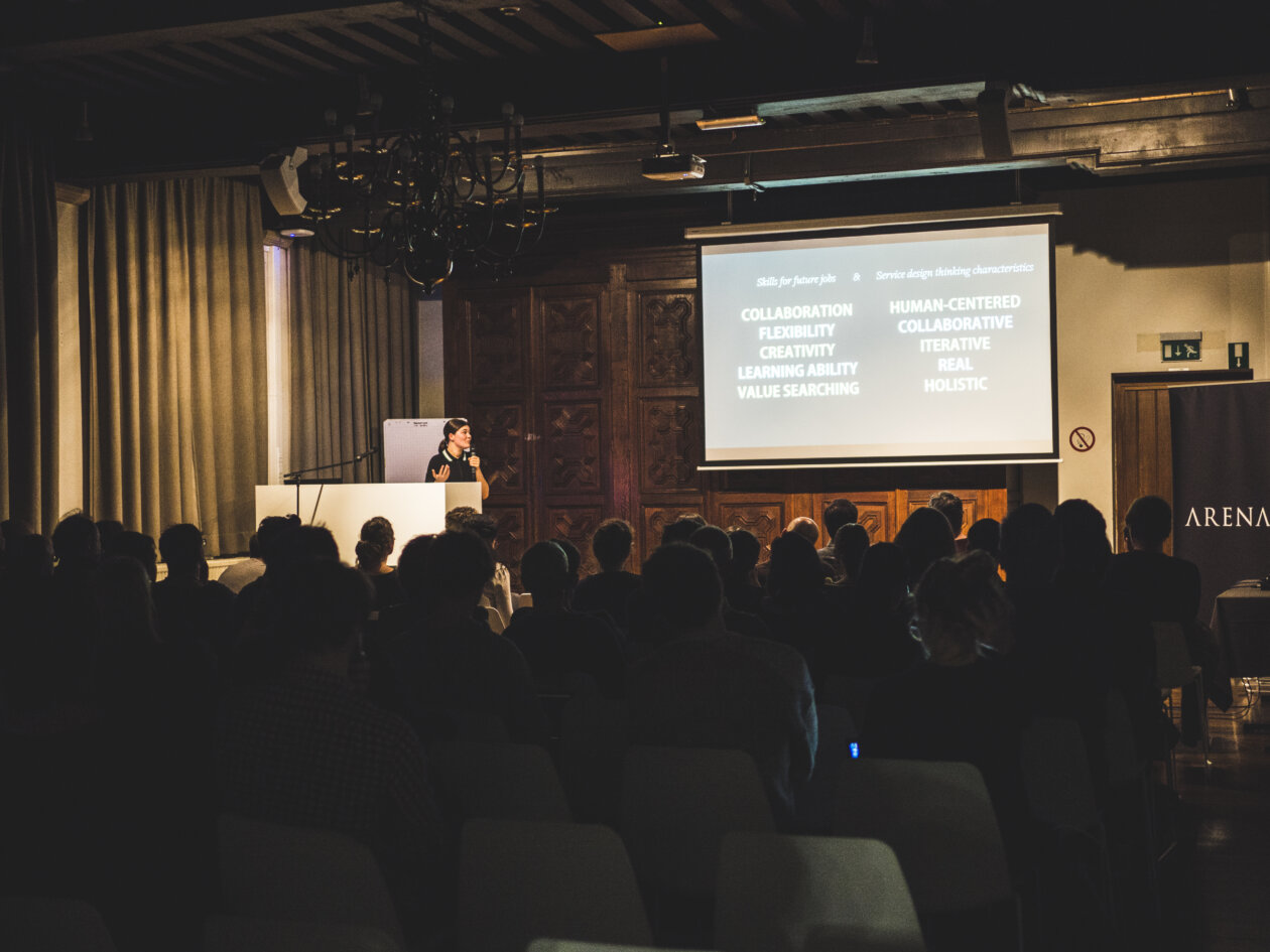 Designexperte spreekt zittend publiek toe met presentatie tijdens ARENA.